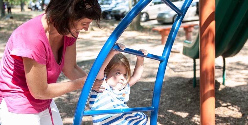 A woman helps a young girl climp up the monkey bar ladder.